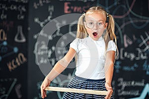 Little blonde girl with a surprised face with a ruler in her hands Pupil girl with big rulers against chalkboard with school