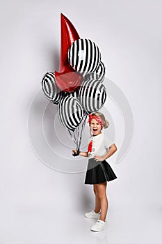 Little blonde girl in t-shirt, red headband, black skirt and sneakers. She holding balloons, posing isolated on white. Full length