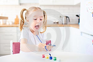 Little blonde girl with two ponytales playing and painting on her hands with blue paint sitting at the light kitchen at home. Self