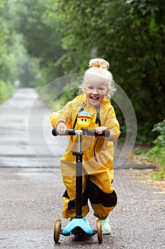 Little blonde girl wearing yellow waterproof overall riding scooter outdoors in countryside. Vertical format