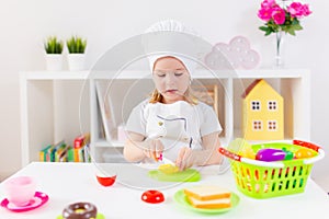 Little blonde girl in white cook uniform playing with toy fruits and vegetables at home, in kindergaten or preschool. Game