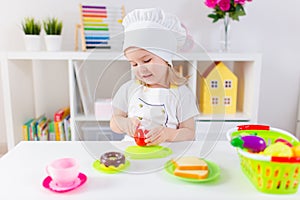 Little blonde girl in white cook uniform playing with toy fruits and vegetables at home, in kindergaten or preschool. Game