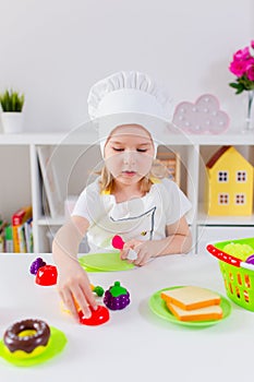 Little blonde girl in white cook uniform playing with toy fruits and vegetables at home, in kindergaten or preschool. Game