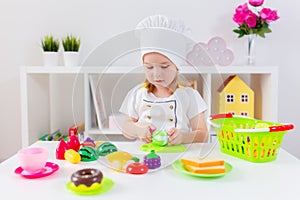 Little blonde girl in white cook uniform playing with toy fruits and vegetables at home, in kindergaten or preschool. Game