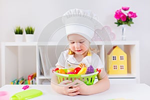 Little blonde girl in white cook uniform playing with toy fruits and vegetables at home, in kindergaten or preschool. Game