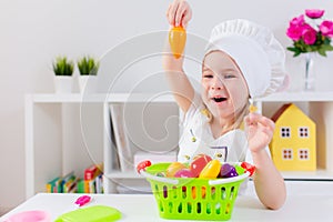 Little blonde girl in white cook uniform playing with toy fruits and vegetables at home, in kindergaten or preschool. Game