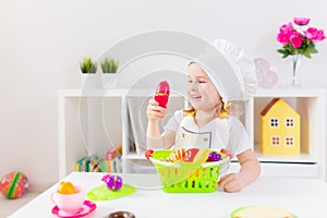 Little blonde girl in white cook uniform playing with toy fruits and vegetables at home, in kindergaten or preschool. Game