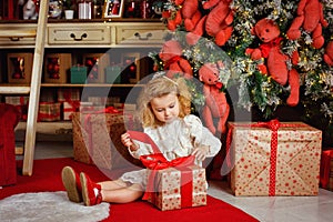 A little blonde girl in a white dress opens a gift sitting near the Christmas tree and lights of garlands