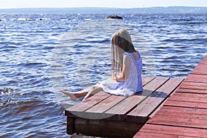 Little blonde girl in a white dress sitting on a wooden pier, surrounded by water
