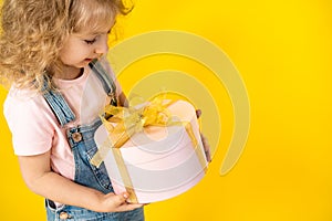 Little blonde girl on a yellow background holds a gift in a pink round box with gold ribbon, copy space