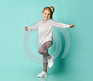 Little blonde kid in shirt with hearts print, checkered pants, white sneakers. Smiling, dancing on blue background. Full length