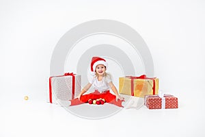 Little blonde kids in Santas hat sitting between gift boxes and playing with christmas balls. Isolated on white background.