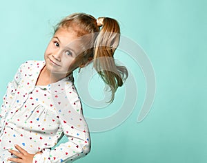 Little blonde schoolgirl with ponytail, in shirt with hearts print. Smiling, hands on hips, posing on blue background. Close up