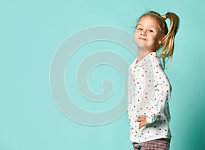 Little blonde schoolgirl with ponytail, in shirt with hearts print. Smiling, hands on hips, posing on blue background. Close up