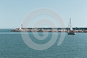 A little boat is sailing in front of the pier of Pesaro harbor with breakwaters and a small red lighthouse Italy, Europe