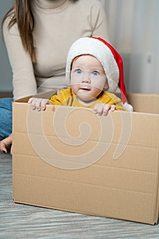 A little boy 1 year old sits in a box wearing a Santa Claus hat on his head and holds on with his hands. Baby mom in the