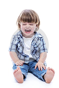 A little boy, 2 years old, sits on the floor and cries. Cute child in jeans and a plaid shirt. Pain and resentment. White