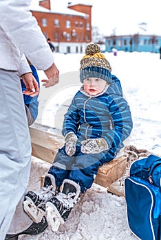 Little boy 3-5 years old, sitting on a bench for shoes, in the winter in a city park on a snow-covered skating rink. In