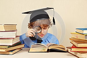 Little boy in academic hat studies an old books with magnifier