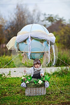 A little boy in a aviator`s hat sits in a basket of a toy balloon in a field