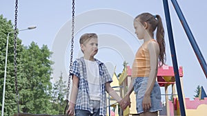 Little boy and beautiful girl with long hair holding hands and talking near the swing, smiling happily. A couple of