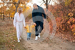 Little boy being holding hands by parents on family autumn walk through countryside. Happy sports family on a walk in