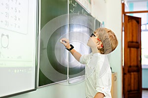 Little boy with big black glasses and white shirt standing near school blackboard with a piece of chalk making smart thinking face