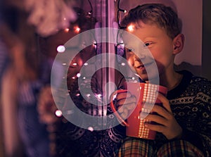 Little boy with big cup of hot drink looks on the window with Christmass Lights