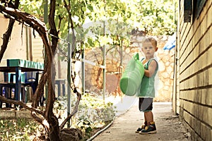 A little boy with a big green watering can is in the yard. Great mother`s helper