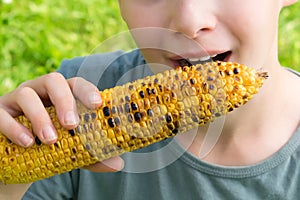 Little boy bites a fried yellow ear of corn sitting on green grass, close-up