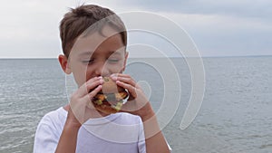 A little boy bites a hamburger. A happy child eats in nature, enjoying a delicious juicy burger. Fast food. A positive