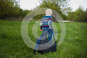 The little boy with blond hair pulling a large backpack chery on the green grass in nature, travel, baby, adventure