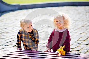 A little boy and a blonde girl near a park bench with yellow leaves in their hands