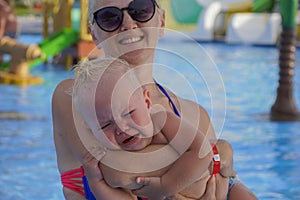 Little boy with blonde hair crying in mom`s arms in a pool