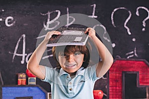 Little boy with a blue t-shirt is holding a black and white notebook to his head in the classroom