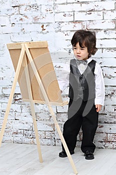 Little boy in bow tie stands next to easel in studio