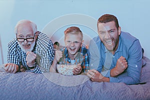 Little boy with bowl of popcorn, his father and grandfather on bed, watching a movie
