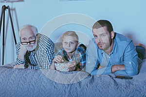 Little boy with bowl of popcorn, his father and grandfather lying on bed and watching