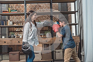 Little boy in boxing gloves kicking a punching pad held by his mother