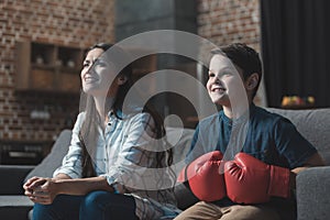 Little boy in boxing gloves sitting on couch with his mother and watching a