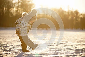 Little boy in bright clothes, hat with pompom and mittens standing in sunshine and playing with snow in sunny snowy park