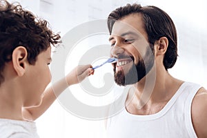 Little boy is brushing teeth of bearded man with toothbrush.