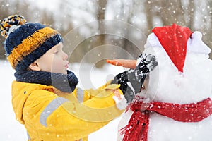 Little boy building funny snowman. Ð¡hild attaches carrot nose to snowman in snowy park