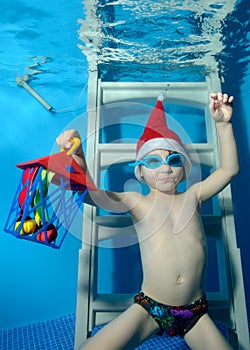 A little boy in a cap Santa Claus with a gift in hand sits underwater on the stairs at the bottom of the pool