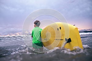 Little boy carries yellow surf board into ocean waves. Surfing First steps concept