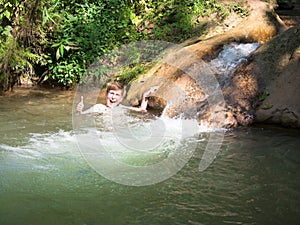 Little boy cheerfully laughs and shows a positive sign with his hand on a background of small bubbling waterfall