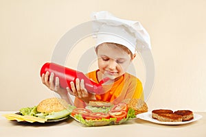 Little boy in chefs hat puts ketchup on hamburger