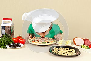 Little boy in chefs hat sniffing cooked pizza