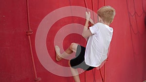 A little boy climbs up ropes with knots on a red wall in an amusement park.