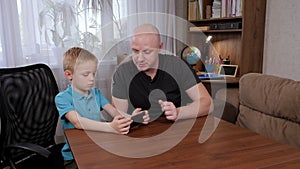 A little boy and dad play video games on the phone at home sitting at the table.